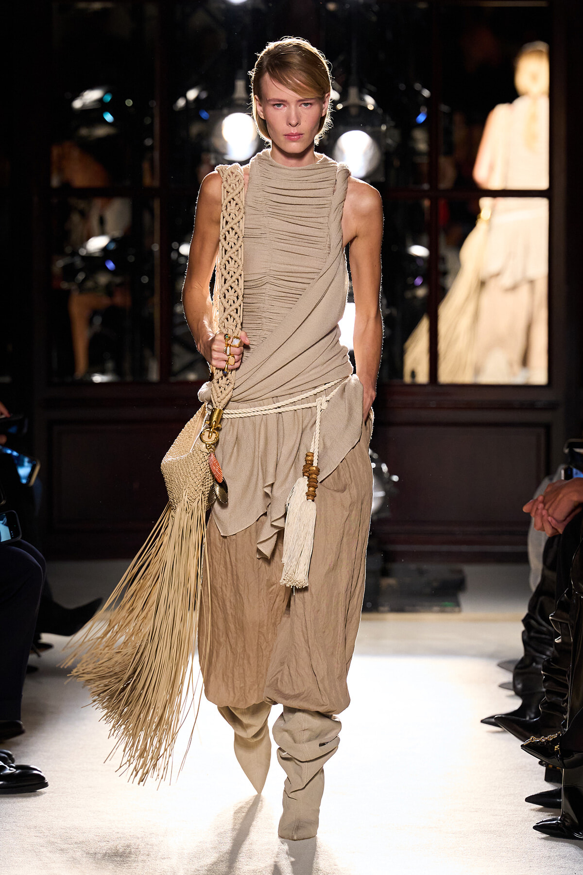 Model walks the runway in a beige draped top and harem-style pants, carrying a long fringed beige bag.