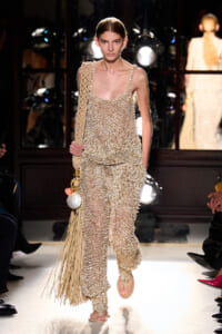 Model walks the runway in a cream crochet jumpsuit with a long-fringe woven bag over her shoulder.
