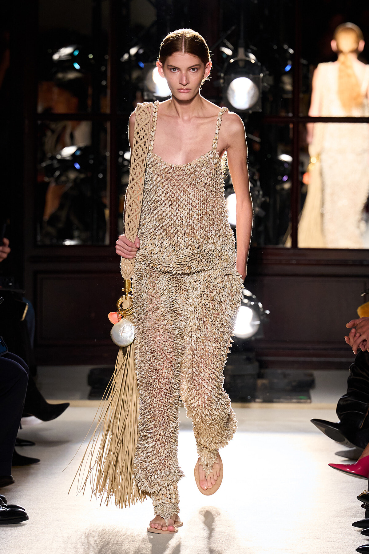 Model walks the runway in a cream crochet jumpsuit with a long-fringe woven bag over her shoulder.