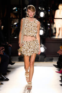 Model walks the runway in a beige shell-embellished crop top and matching mini skirt, carrying a knit bag with shell accents and wearing layered sandals.