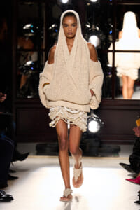 Model walking a runway in a chunky beige knit hooded sweater with exposed shoulders and a layered fringed mini skirt, barefoot sandals in a neutral show look.