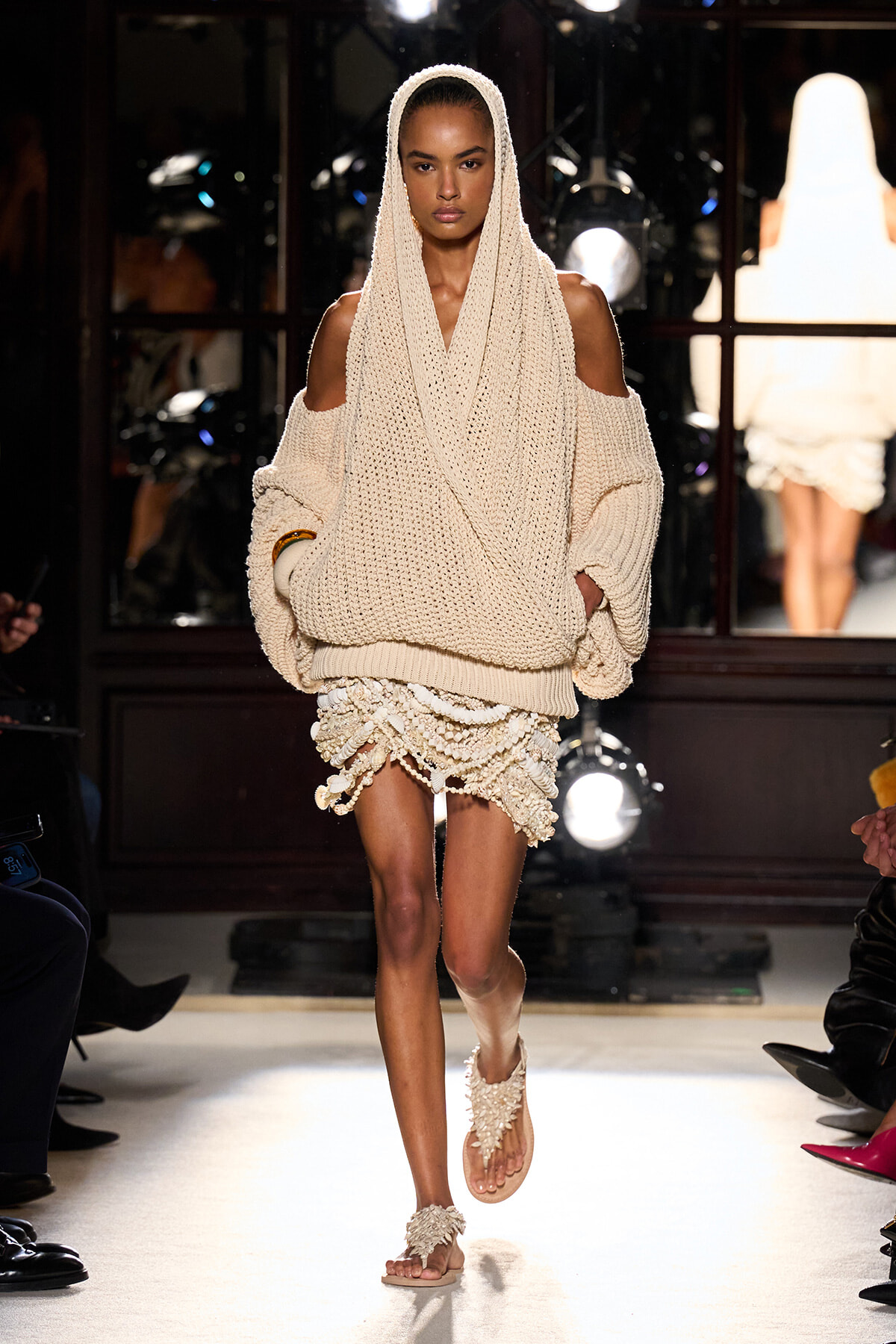 Model walking a runway in a chunky beige knit hooded sweater with exposed shoulders and a layered fringed mini skirt, barefoot sandals in a neutral show look.
