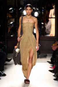 Model walks the runway in a gold strapless knit gown with a high slit, carrying a fringed brown bag adorned with shells.