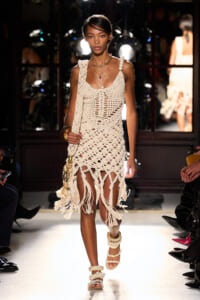Model walks a runway in a cream crochet dress with fringe and layered necklaces, carrying a tassel bag and wearing chunky sandals.