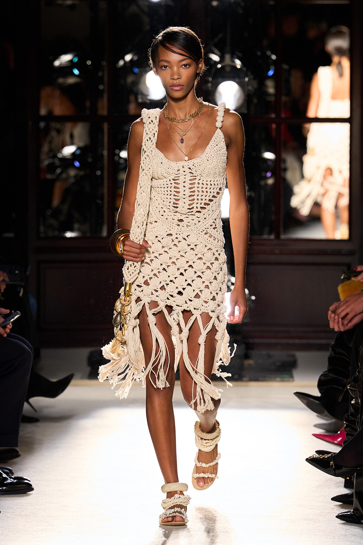 Model walks a runway in a cream crochet dress with fringe and layered necklaces, carrying a tassel bag and wearing chunky sandals.