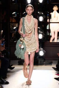 Model on runway wearing a cream beaded fringe dress with layered necklaces, carrying a teal fringed bag.