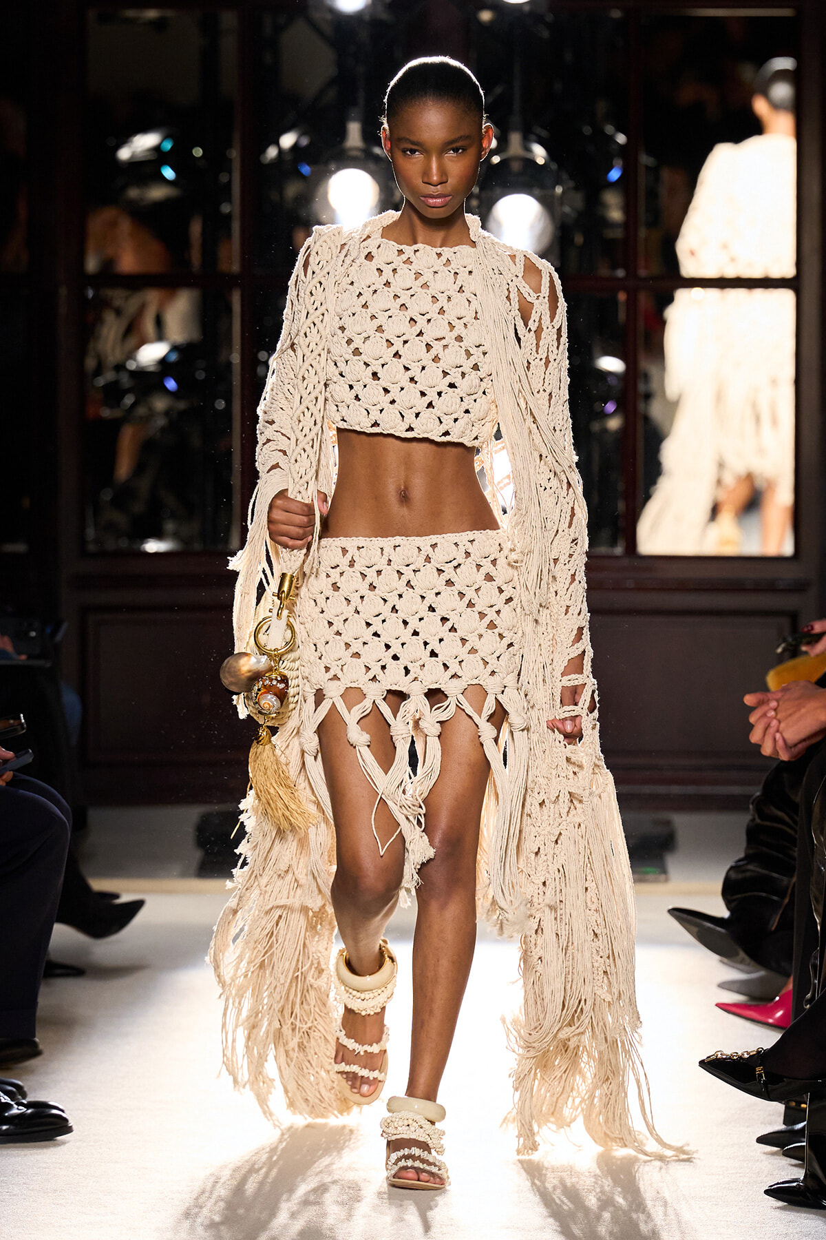 Female model walks a runway in a cream crochet crop top and matching fringe skirt, carrying a tassel-adorned bag.