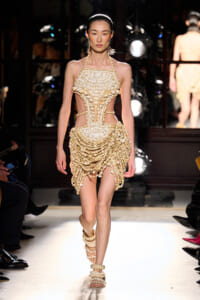 Model walks the runway in a strapless beige beaded gown with shell-like embellishments and cutouts at the waist.