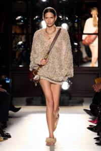 Model walking the runway in a chunky beige knit sweater dress with a brown crossbody bag and spiky beige sandals, accessorized with layered gold necklaces.