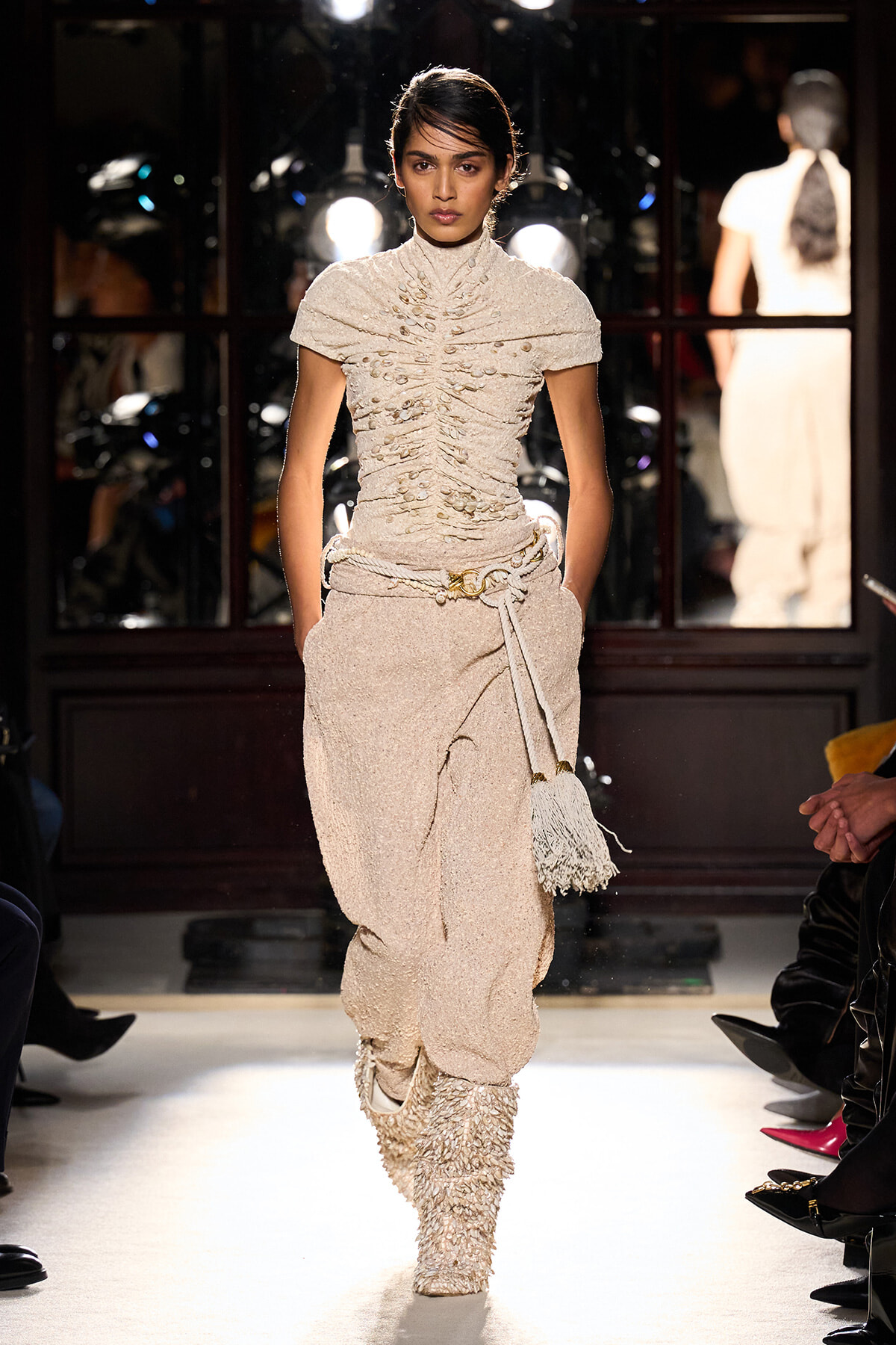Model walks the runway in a beige textured high-neck top with ruched detailing, matching belted trousers, and a fringed tassel bag.