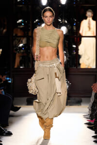Model walking the runway in a tan draped one-shoulder crop top, oversized beige harem pants, and tan suede boots with a macrame bag and tassels slung over the shoulder.