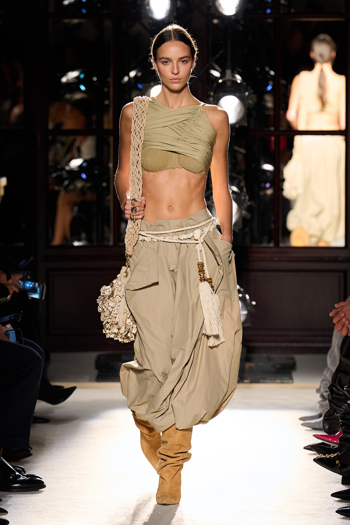 Model walking the runway in a tan draped one-shoulder crop top, oversized beige harem pants, and tan suede boots with a macrame bag and tassels slung over the shoulder.