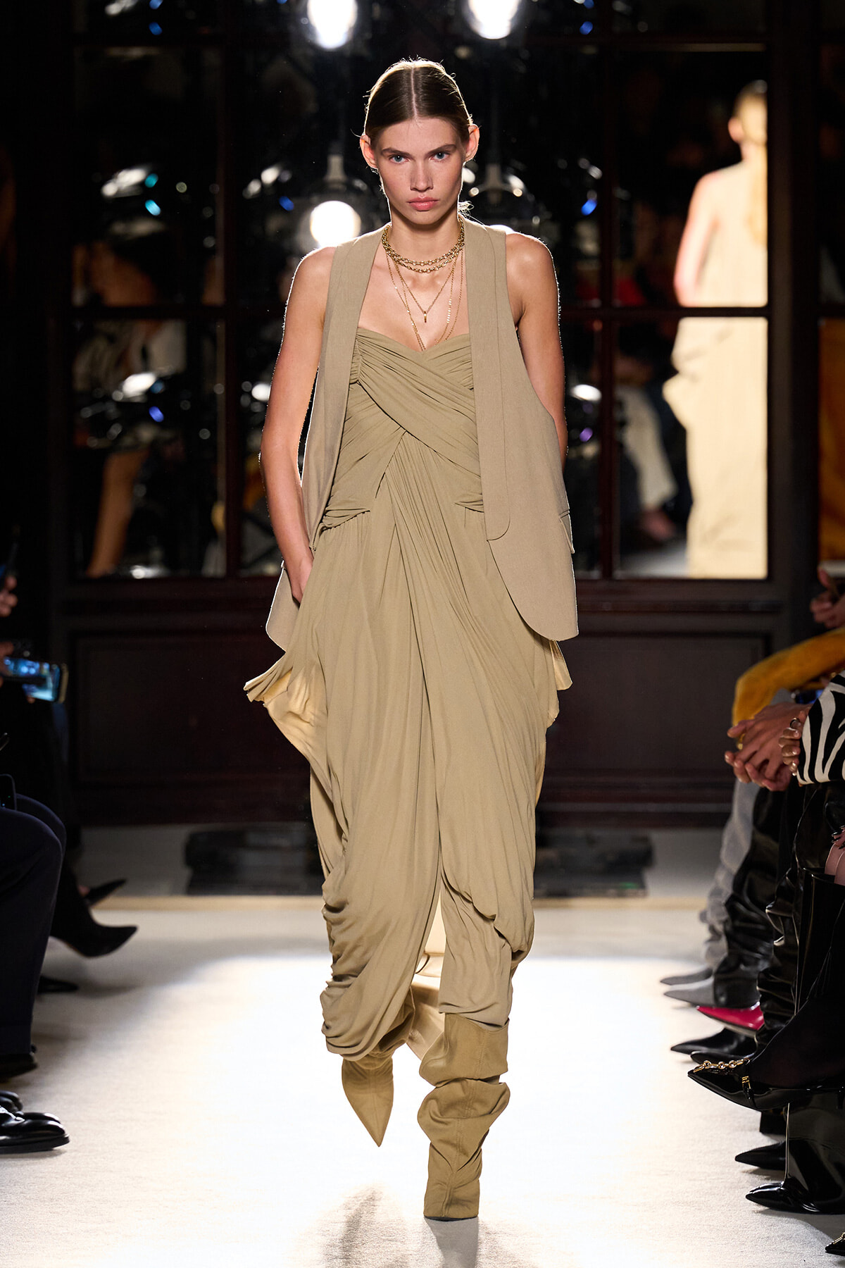 Model walking a beige draped gown with a long sleeveless vest and layered necklaces on a runway.