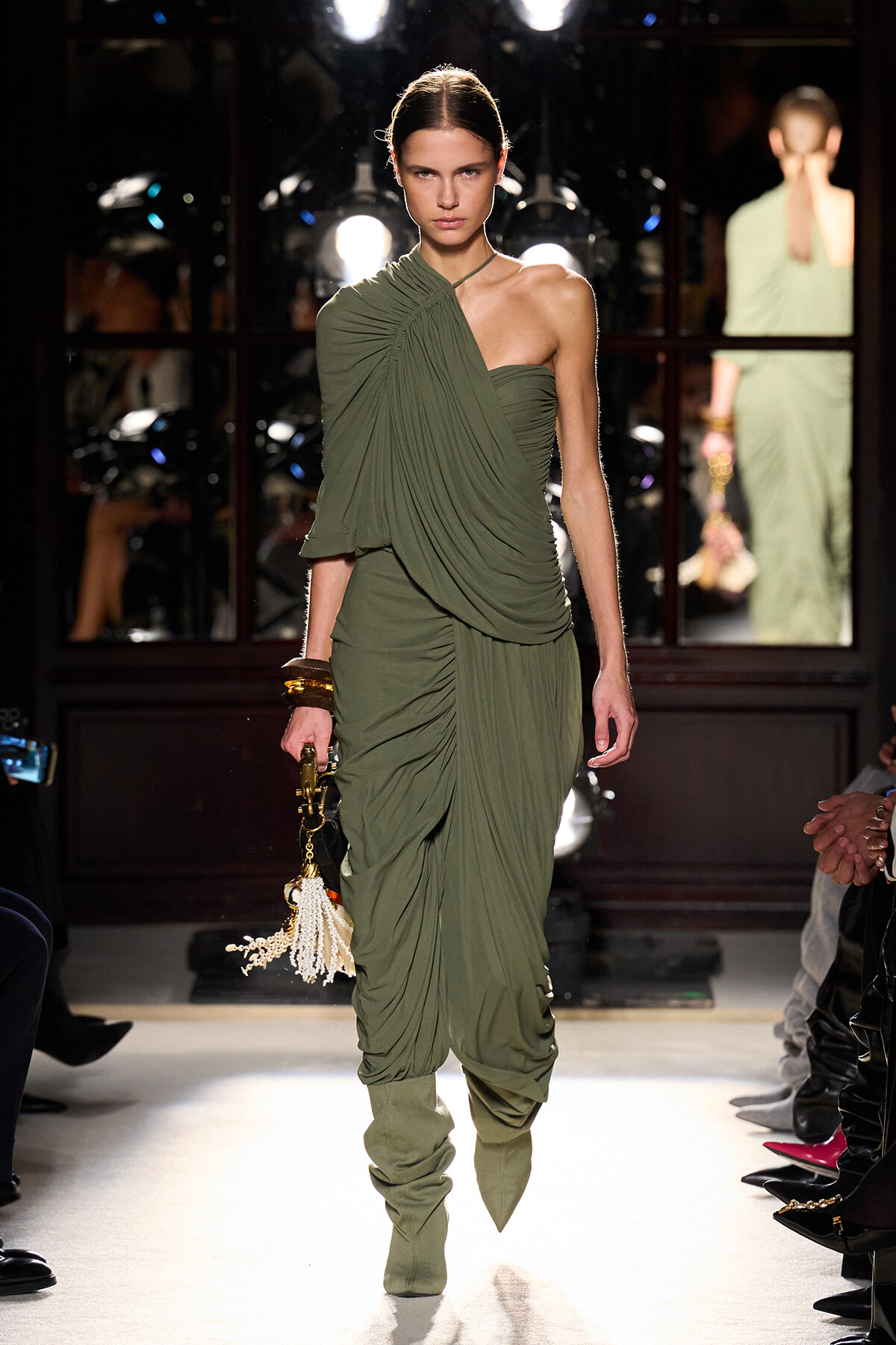 Model walking runway in olive green one-shoulder draped dress with ruched details and matching slouchy boots, carrying a tassel-tied handbag.