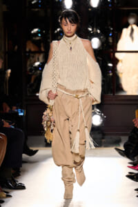 Model walking a runway in a cream open-shoulder knit sweater and tan harem-style pants with tassel accents and a decorative bag