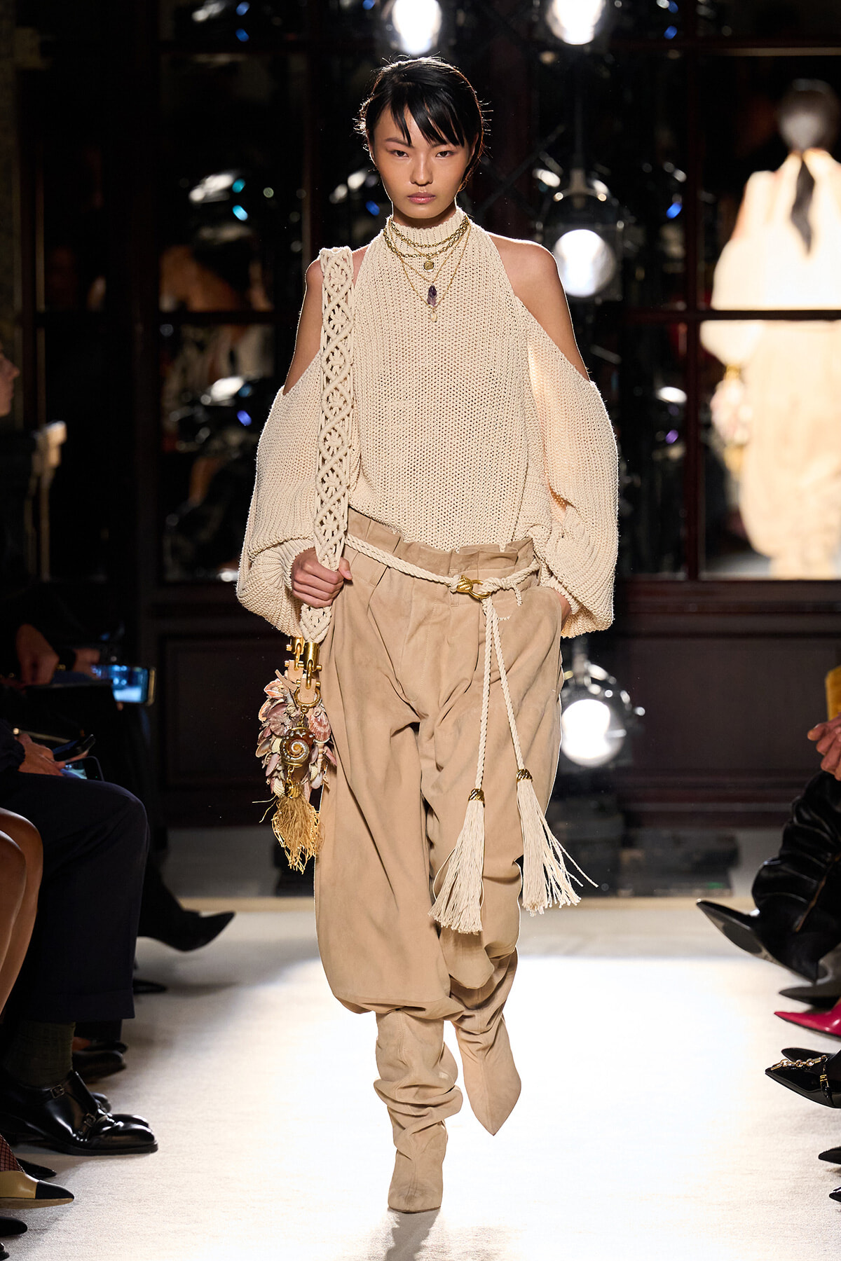 Model walking a runway in a cream open-shoulder knit sweater and tan harem-style pants with tassel accents and a decorative bag