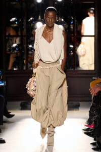 Model walks a beige, layered outfit on the runway: cream draped knit top with voluminous beige pants and a shell‑embellished bag.