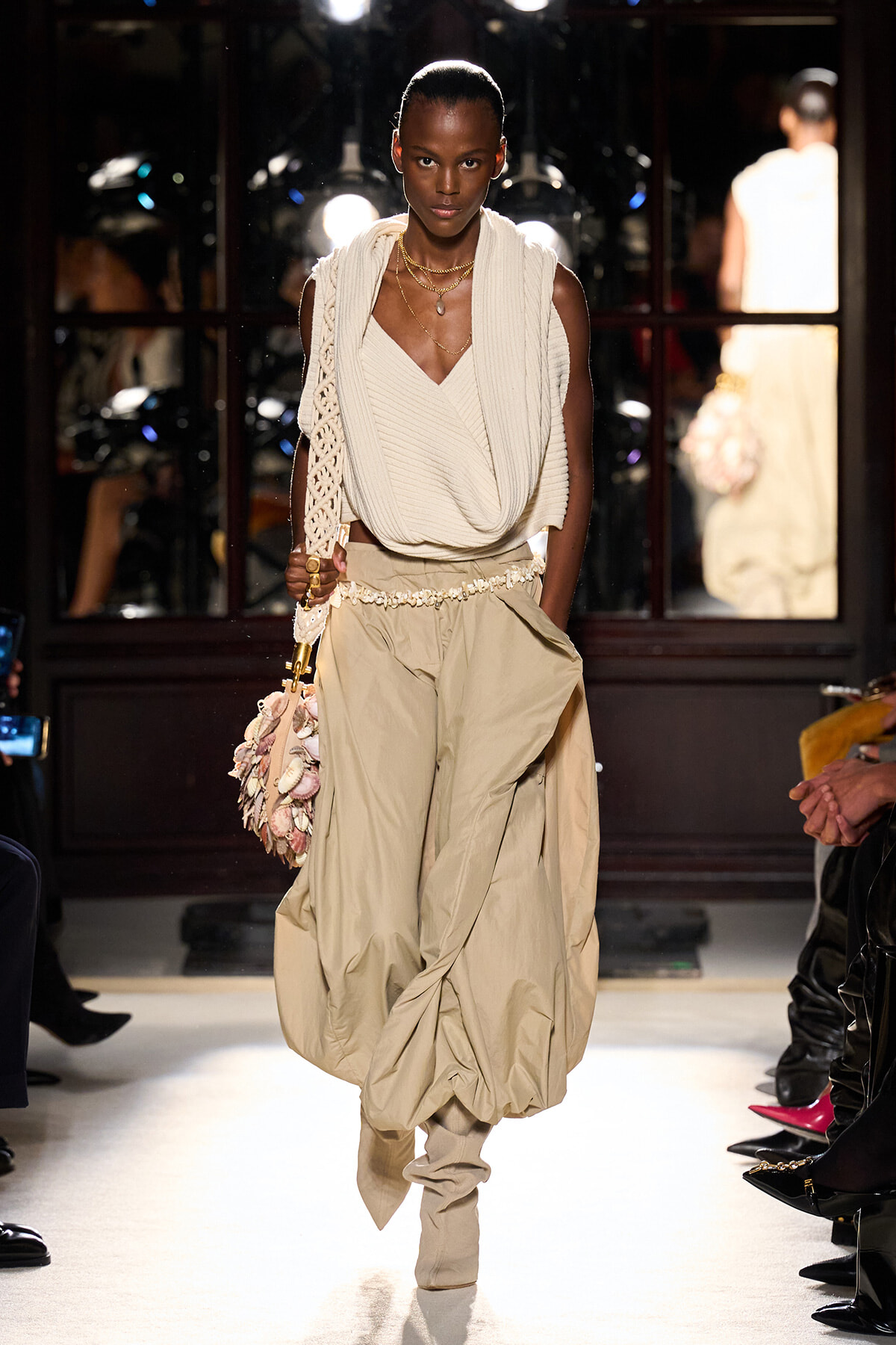 Model walks a beige, layered outfit on the runway: cream draped knit top with voluminous beige pants and a shell‑embellished bag.