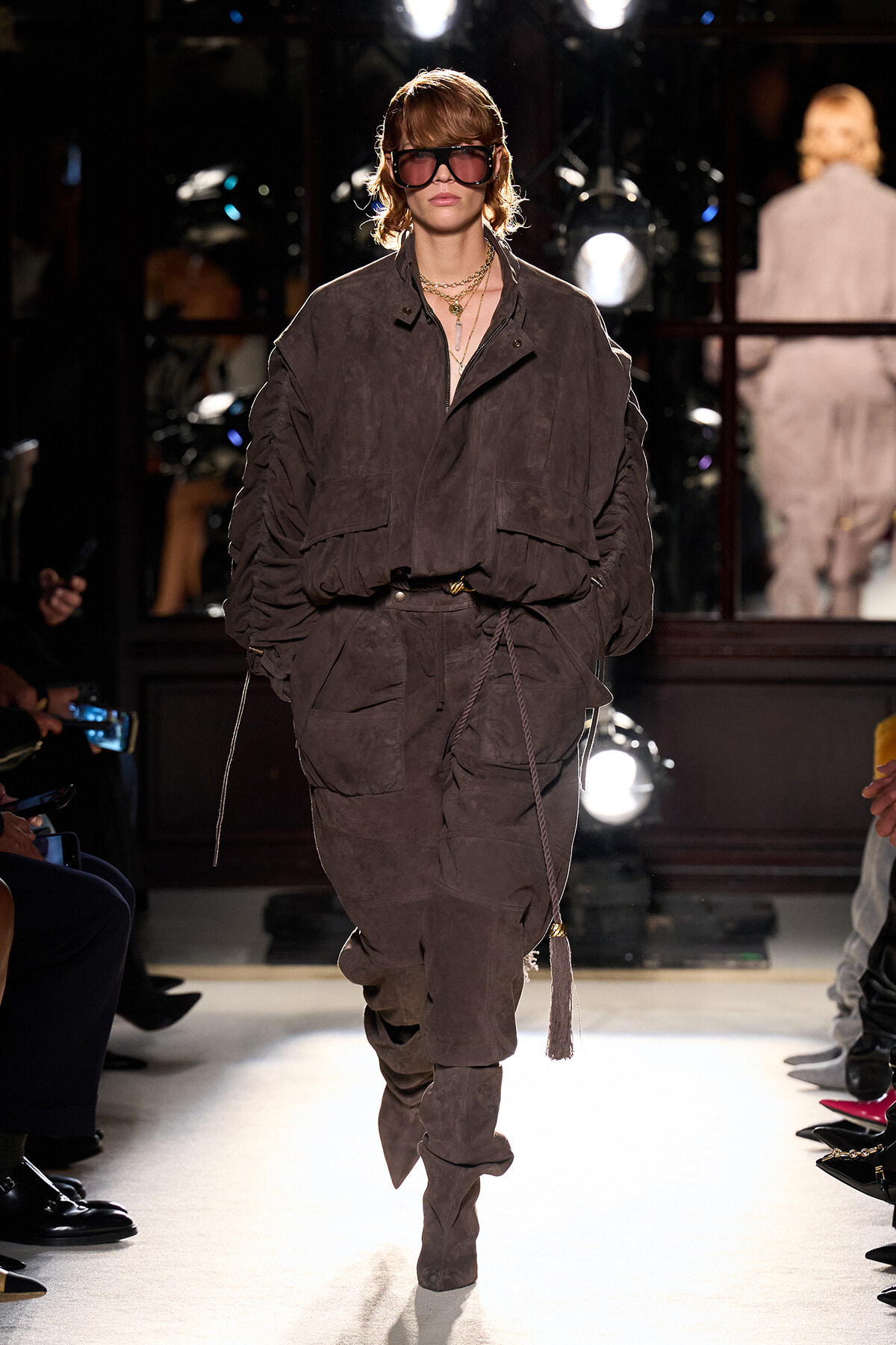 Model walks a runway in a loose brown suede utility suit with large pockets, matching pants, and chunky sunglasses, accessorized with layered necklaces.