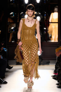 Model walks the runway in a tan crochet dress with fringe and layered necklaces, carrying a fringed bag.
