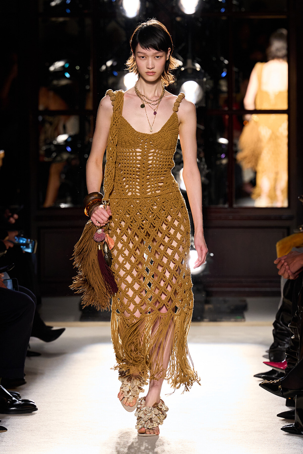 Model walks the runway in a tan crochet dress with fringe and layered necklaces, carrying a fringed bag.