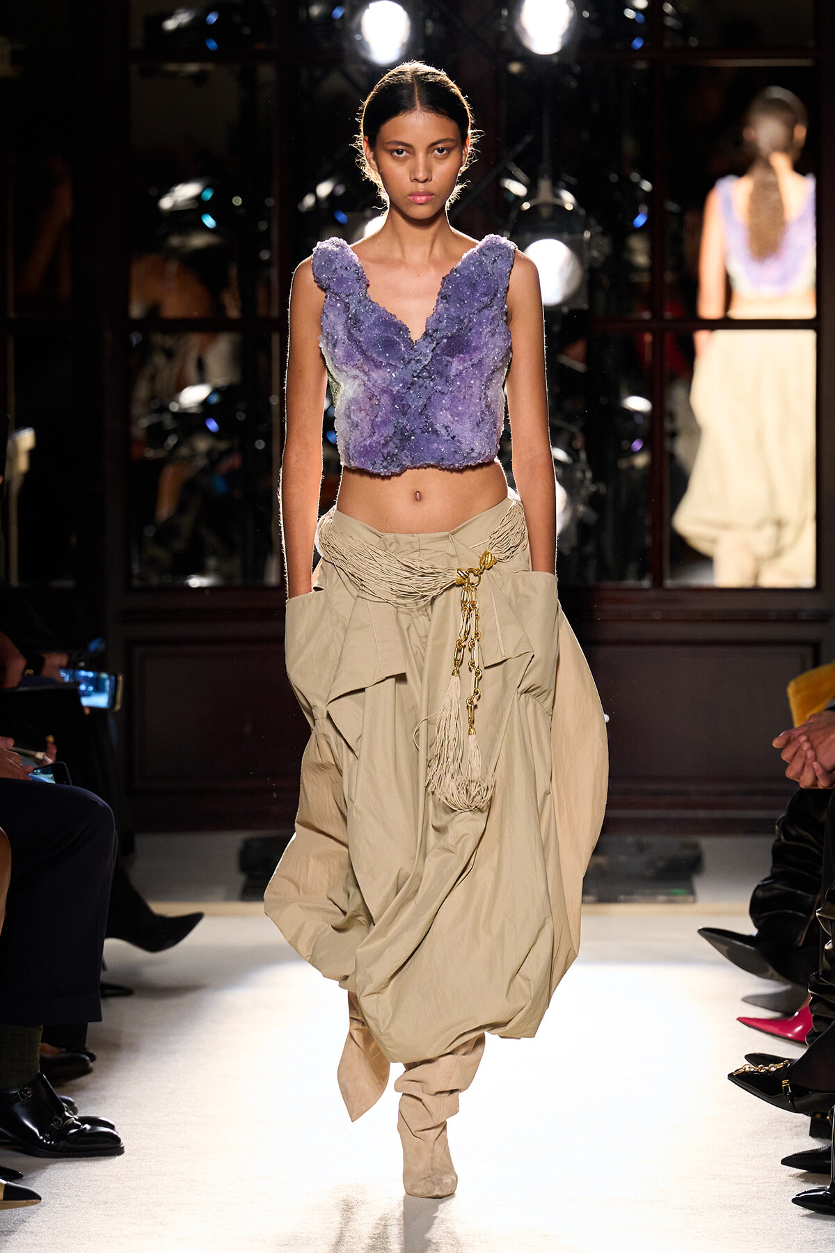 Model walks a runway wearing a lavender textured crop top and voluminous beige draped skirt with a rope belt and boots.