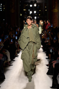 Model walks a fashion runway in an oversized olive-green utility outfit with a rope belt and tassels, audience capturing photos on phones on both sides.