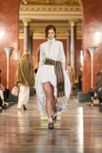 Model walking a fashion runway in a white blouse and dramatic balloon skirt with a striped brown belt/scarf, in a grand ornate hall setting.