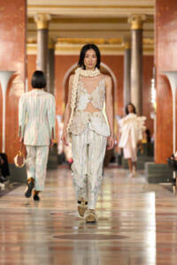 Model walks a luxurious runway in a cream lace bodice with sheer panels and loose floral-striped pants, accessorized with a chunky scarf around the neck.
