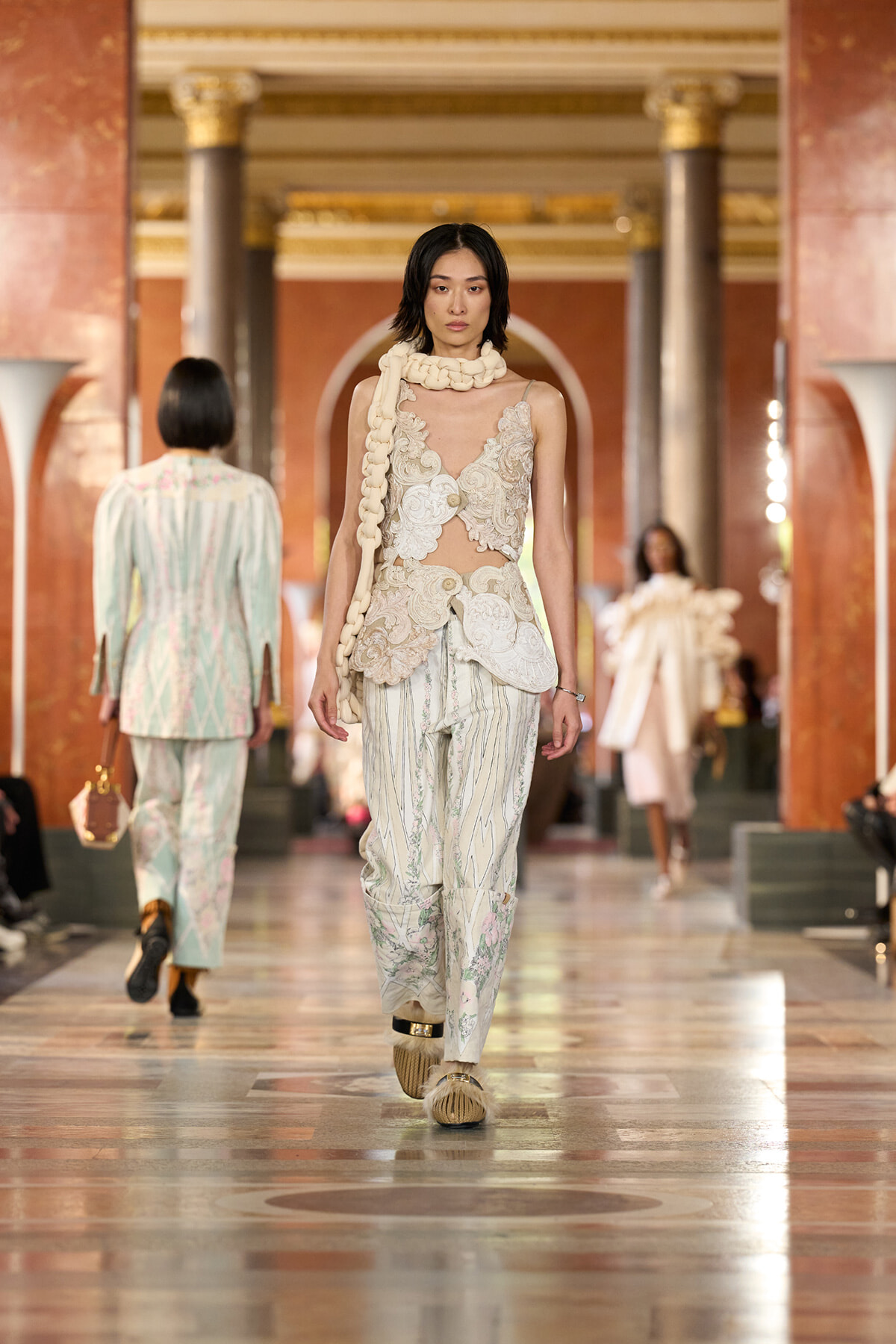 Model walks a luxurious runway in a cream lace bodice with sheer panels and loose floral-striped pants, accessorized with a chunky scarf around the neck.