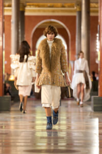 Model walks a runway in a beige embroidered coat with a large fur collar, white layered pants, blue ankle boots, and a small handbag.
