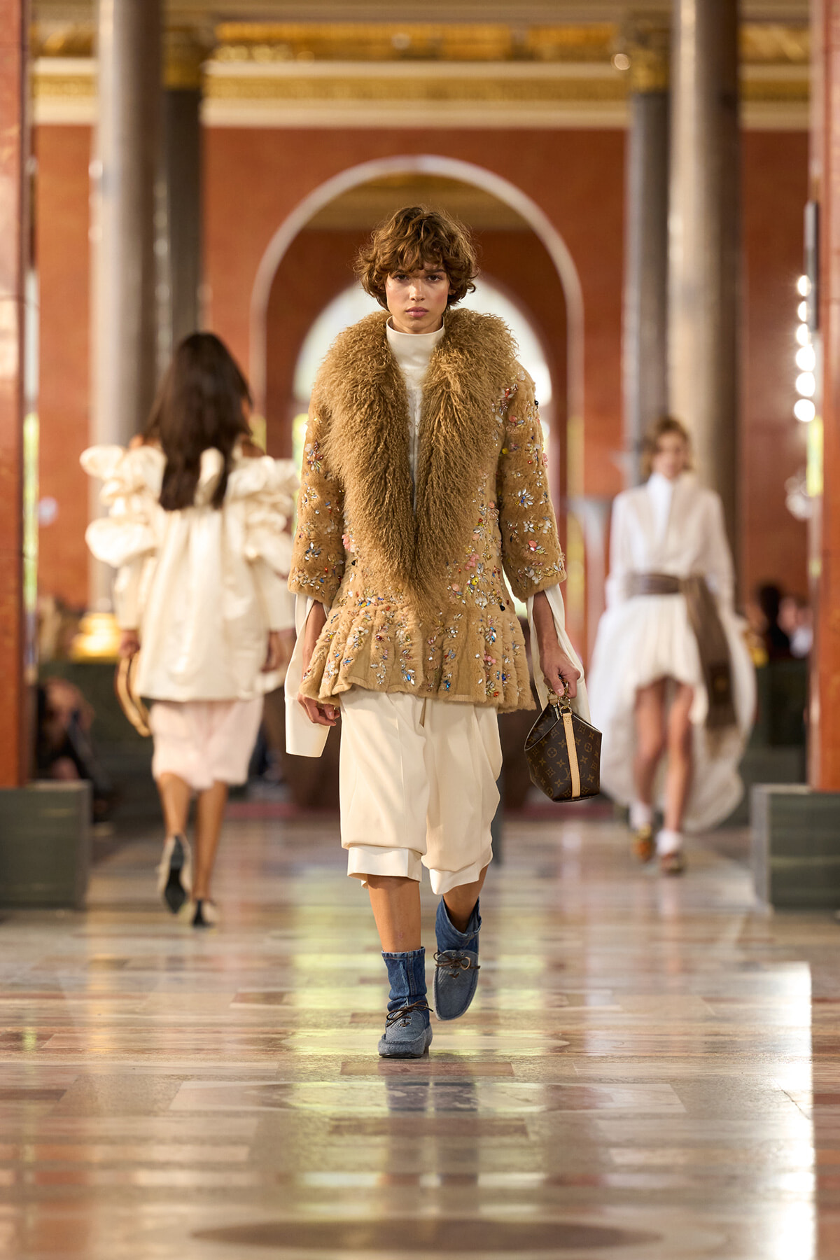 Model walks a runway in a beige embroidered coat with a large fur collar, white layered pants, blue ankle boots, and a small handbag.