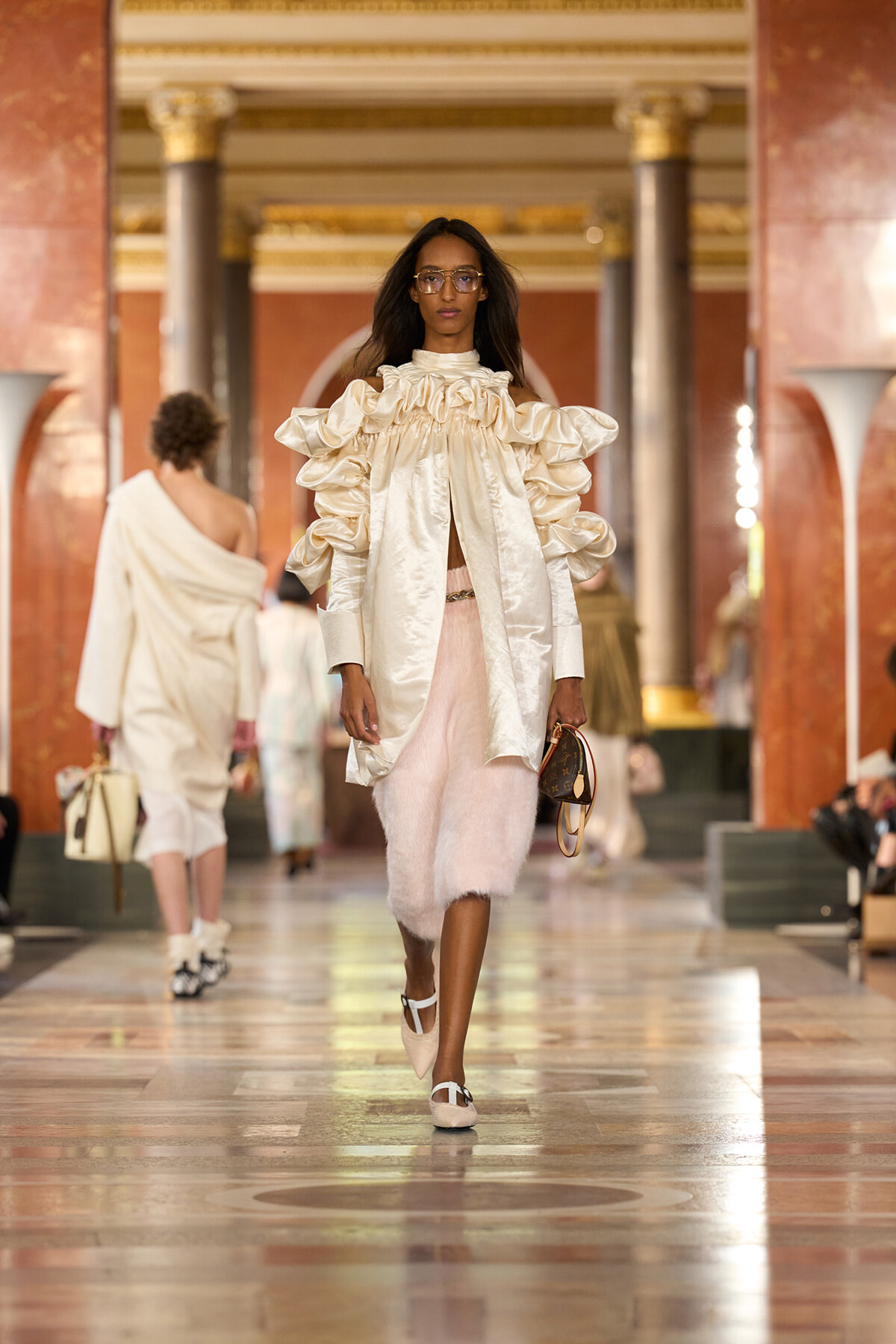 Female model walks a runway in a cream satin ruffled cape-like top and pale pink fuzzy midi skirt, holding a small round handbag in a grand hall setting.