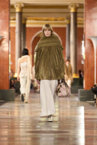 Model walking a fashion runway in an oversized olive green poncho with turtleneck, white wide pants, and a patterned handbag in a grand hall setting.