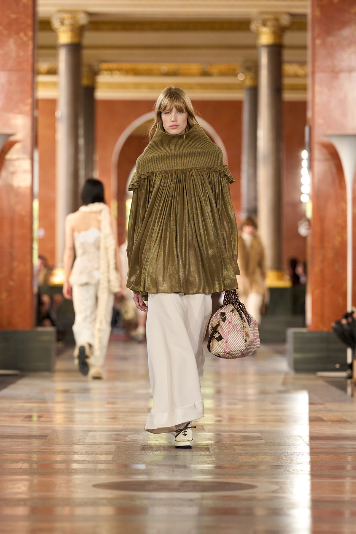 Model walking a fashion runway in an oversized olive green poncho with turtleneck, white wide pants, and a patterned handbag in a grand hall setting.
