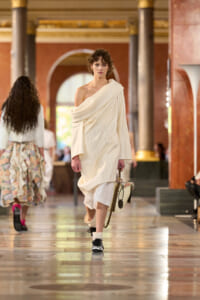 Model walking a fashion runway in a cream draped one-shoulder dress, carrying a beige handbag in an ornate indoor venue with columns and arches.