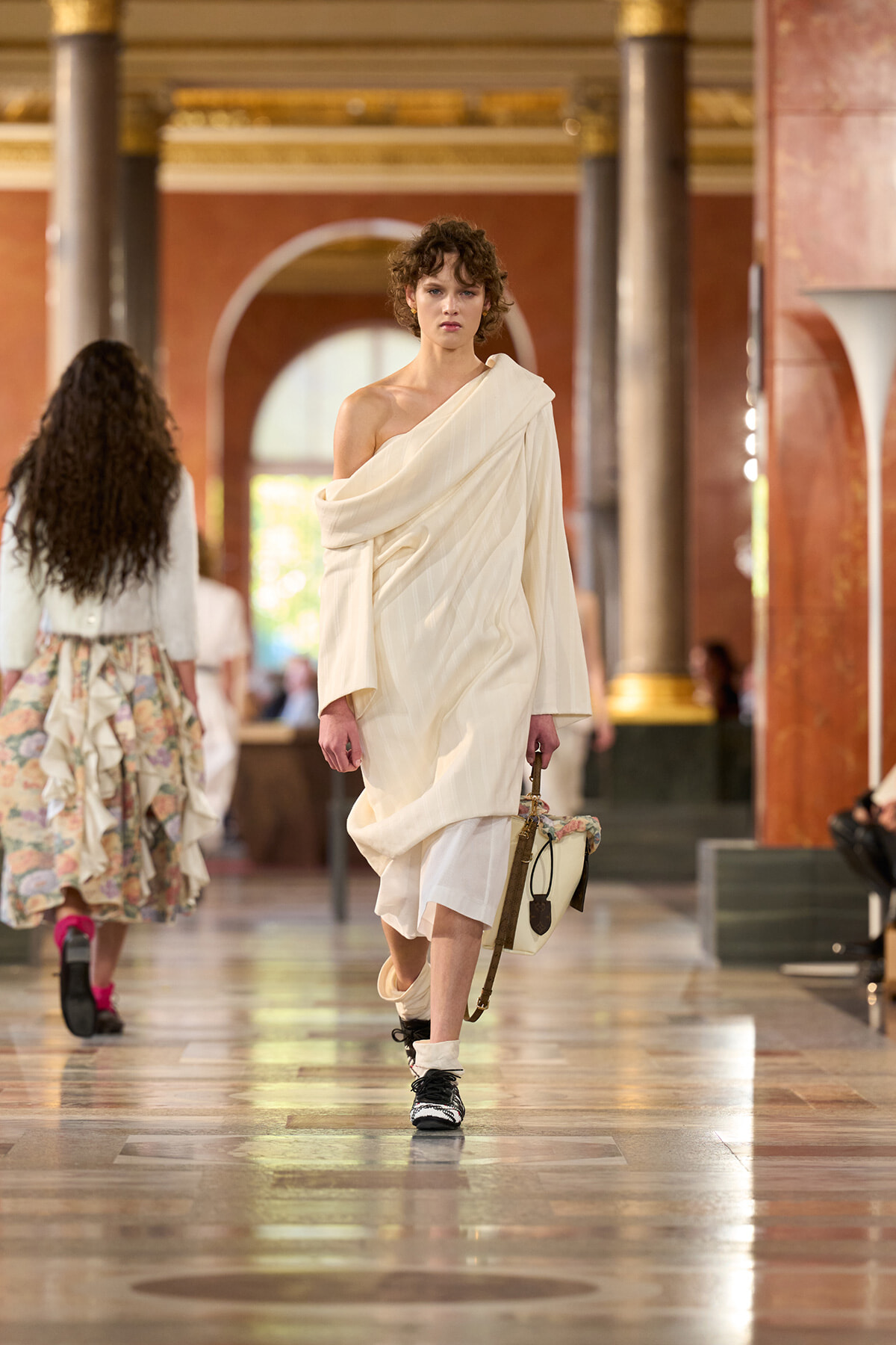 Model walking a fashion runway in a cream draped one-shoulder dress, carrying a beige handbag in an ornate indoor venue with columns and arches.