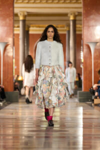 Model walks a runway in a pale knit cardigan and a layered, floral-printed ruffle skirt inside an ornate hall.