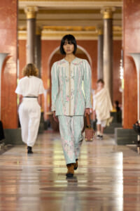 Model walks a fashion show in a pastel mint-green suit with pink floral embroidery, holding a brown handbag.