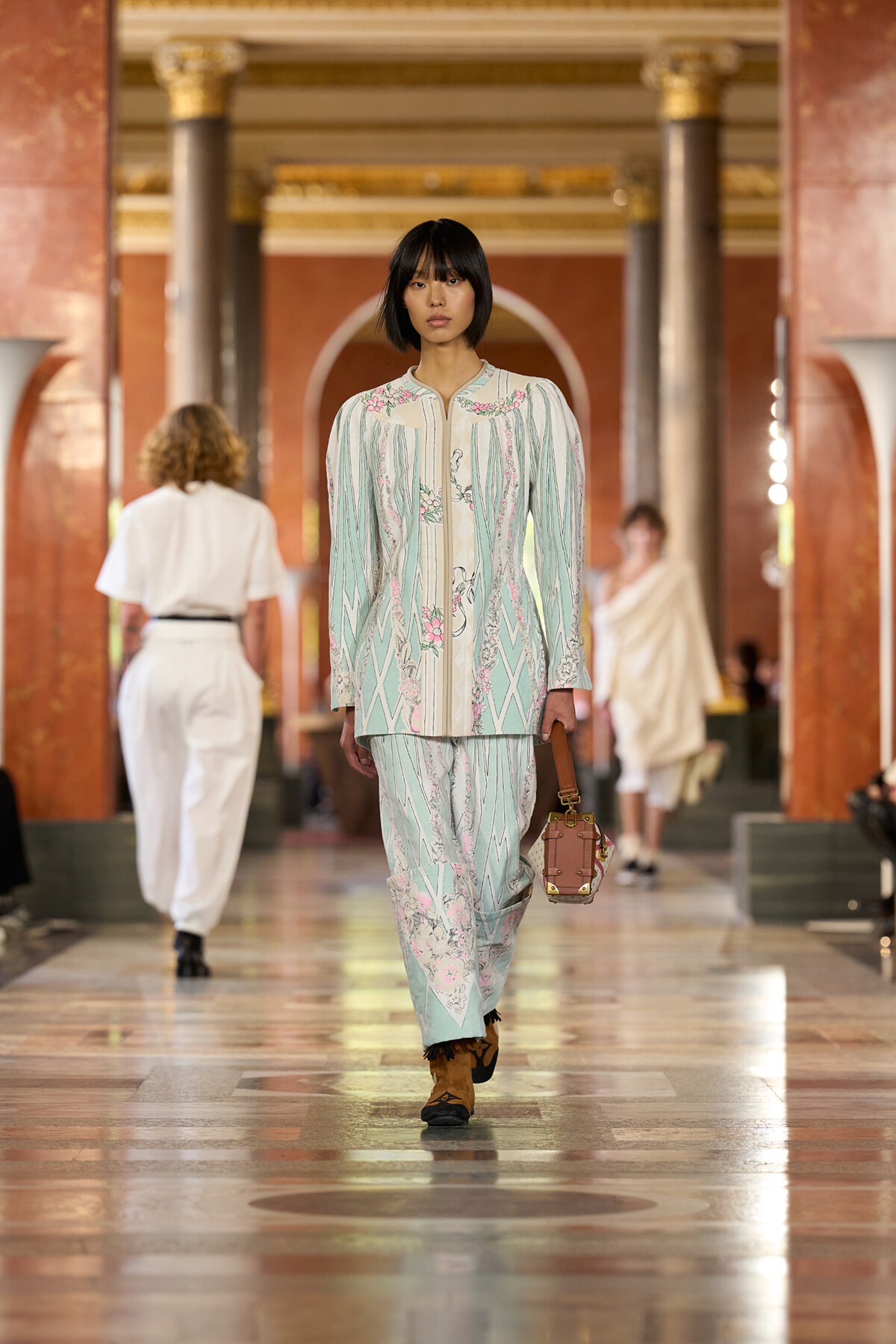 Model walks a fashion show in a pastel mint-green suit with pink floral embroidery, holding a brown handbag.
