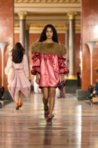 Model walking a fashion runway in a pink satin mini dress with a brown fur collar inside a marble hall.