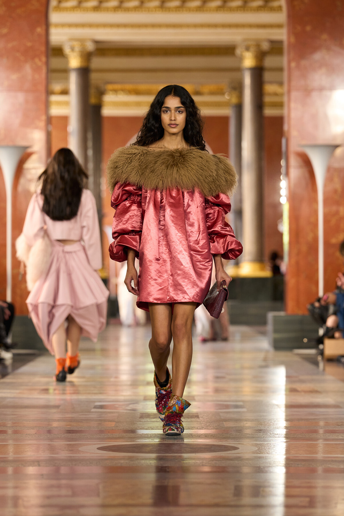 Model walking a fashion runway in a pink satin mini dress with a brown fur collar inside a marble hall.