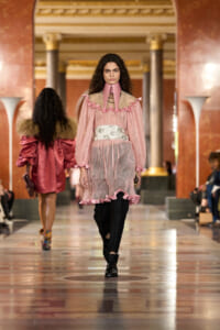 Model walks the runway in a pink sheer dress with ruffled trim over black pants, in a grand marble hall.