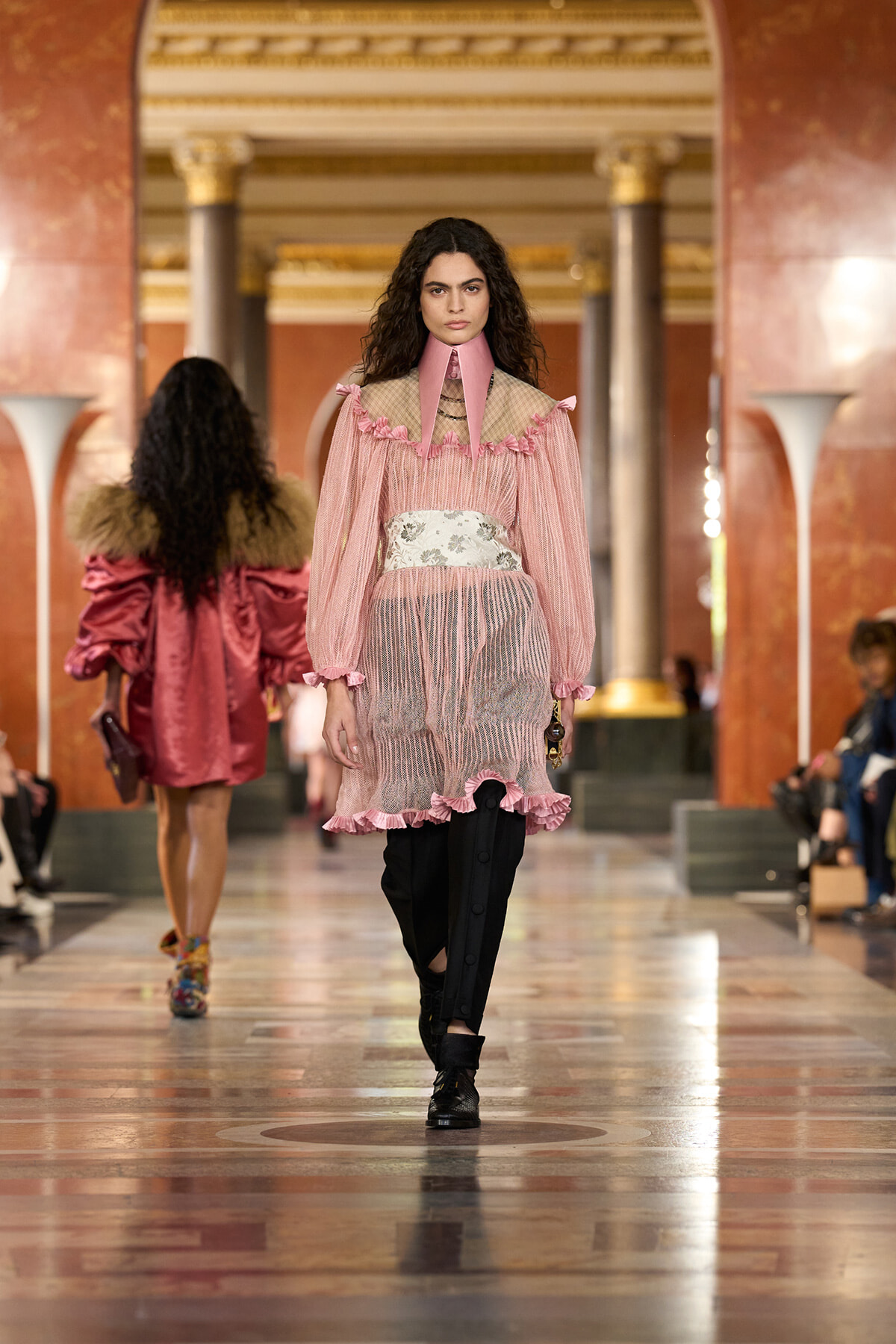 Model walks the runway in a pink sheer dress with ruffled trim over black pants, in a grand marble hall.
