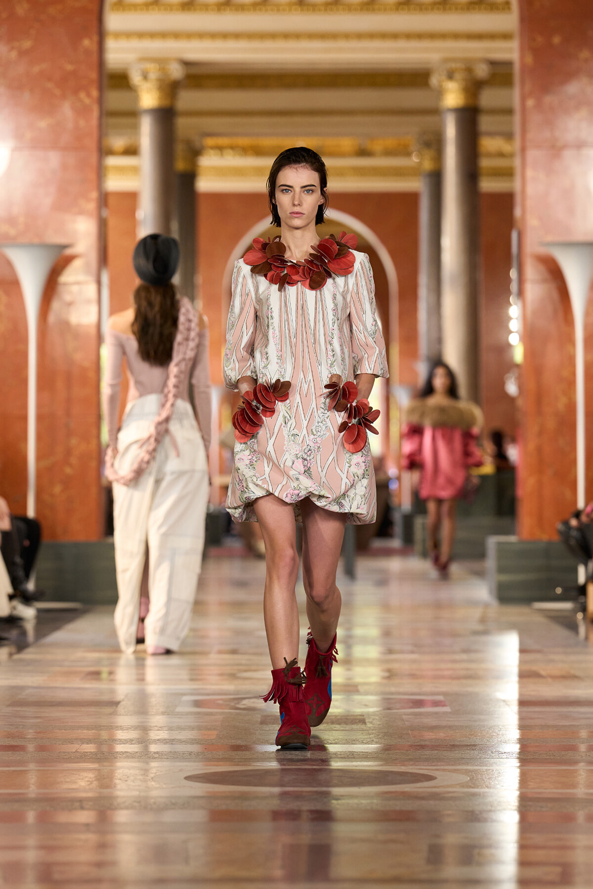 Runway model in a pale pink patterned dress with large red flower appliqués at the neckline and hips, paired with red fringe boots, in an ornate hall.