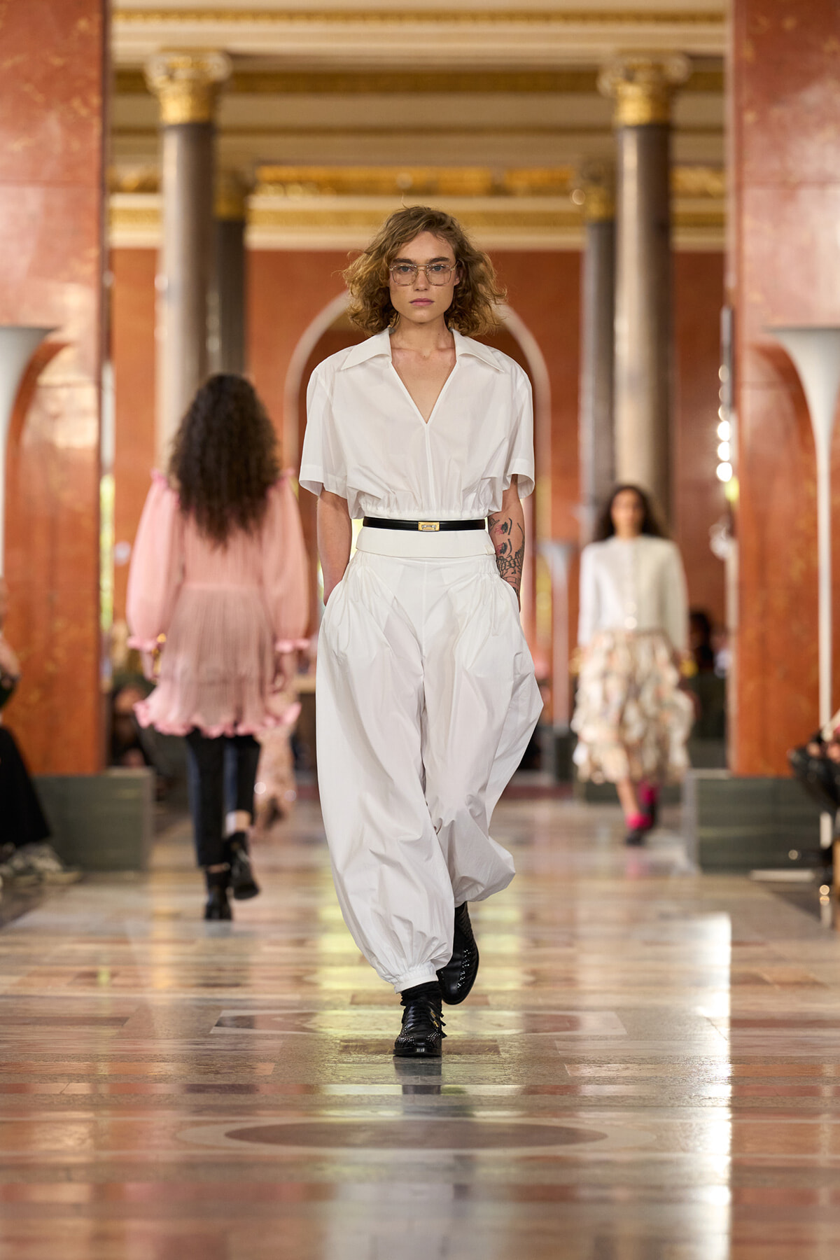 Runway model in a white blouse and oversized white pants with a black belt walking down a marble hall runway.