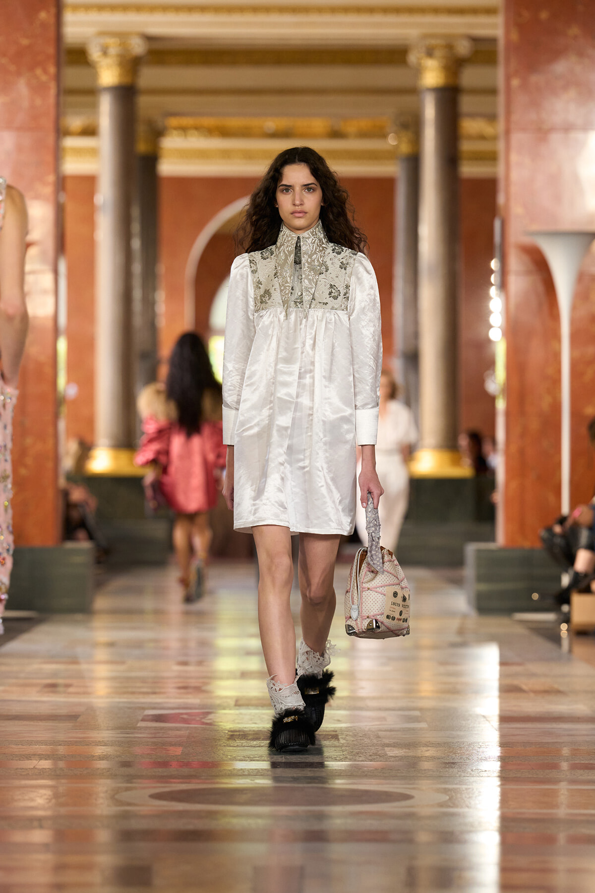 Model walks a fashion runway in a white, knee-length dress with an ornate lace yoke in a grand marble hall.