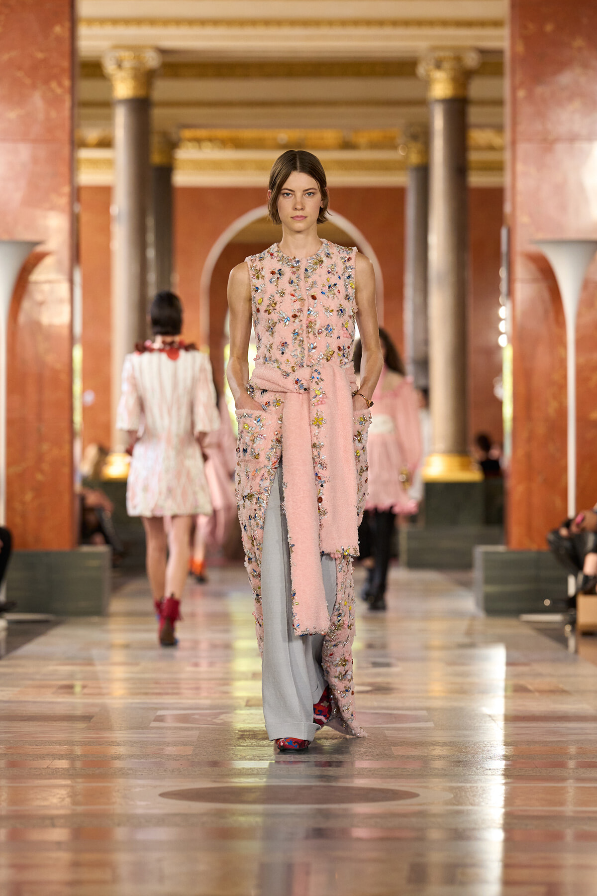 Model walks a fashion runway in a pale pink, sleeveless, floral-embellished jumpsuit with a matching scarf tied around the waist in a grand columned hall.