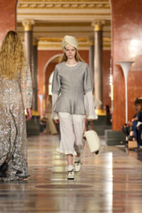 Model on a runway wearing a gray peplum top, white blouse, ivory cropped pants, cream turban, and a small cream handbag.