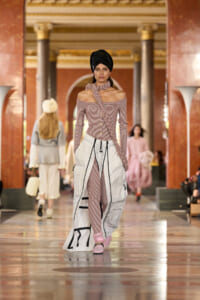 Model walks a runway in a gold-and-marble hallway wearing a striped off-shoulder top, matching leggings, and a white multi-pocket coat.